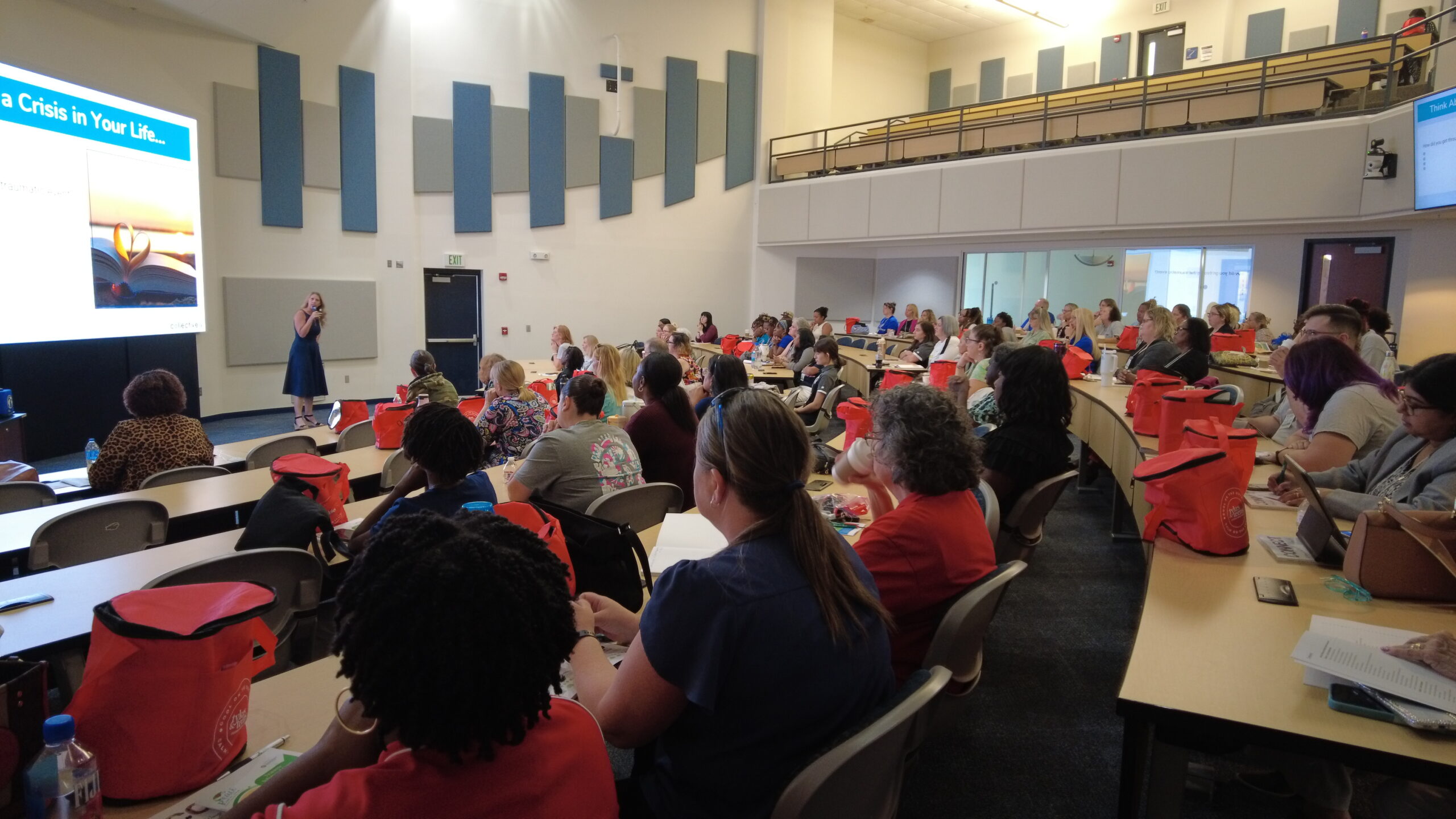 A speaker presents to a full audience in a large lecture hall during the Trauma-Informed Care Symposium. Attendees sit at curved rows of desks, taking notes and listening attentively, with presentation slides projected at the front of the room.