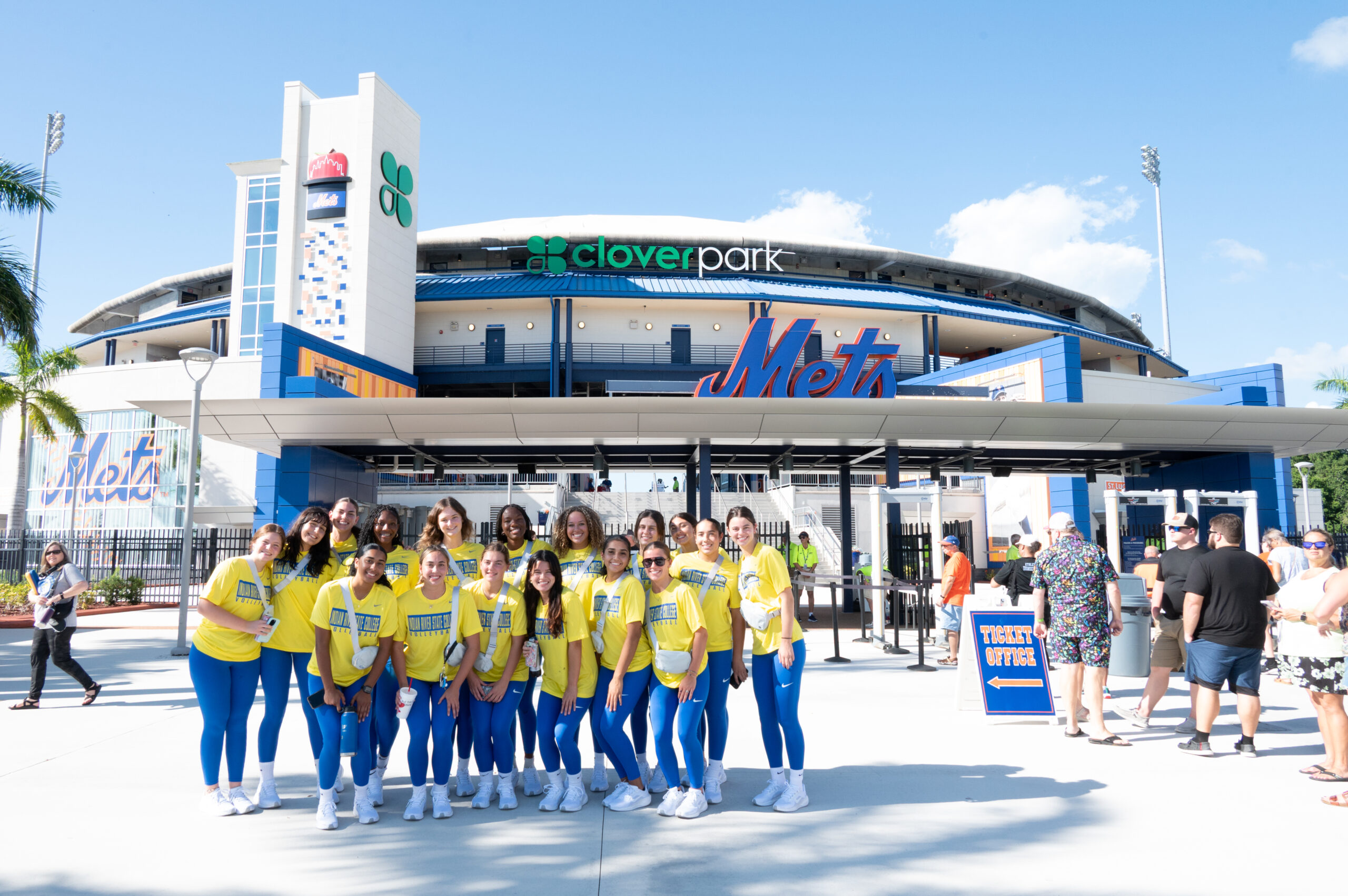 A group of students in matching yellow shirts and blue pants pose for a photo outside Clover Park, home of the St. Lucie Mets. The stadium and its large Mets logo are visible in the background, with other visitors entering the gates.