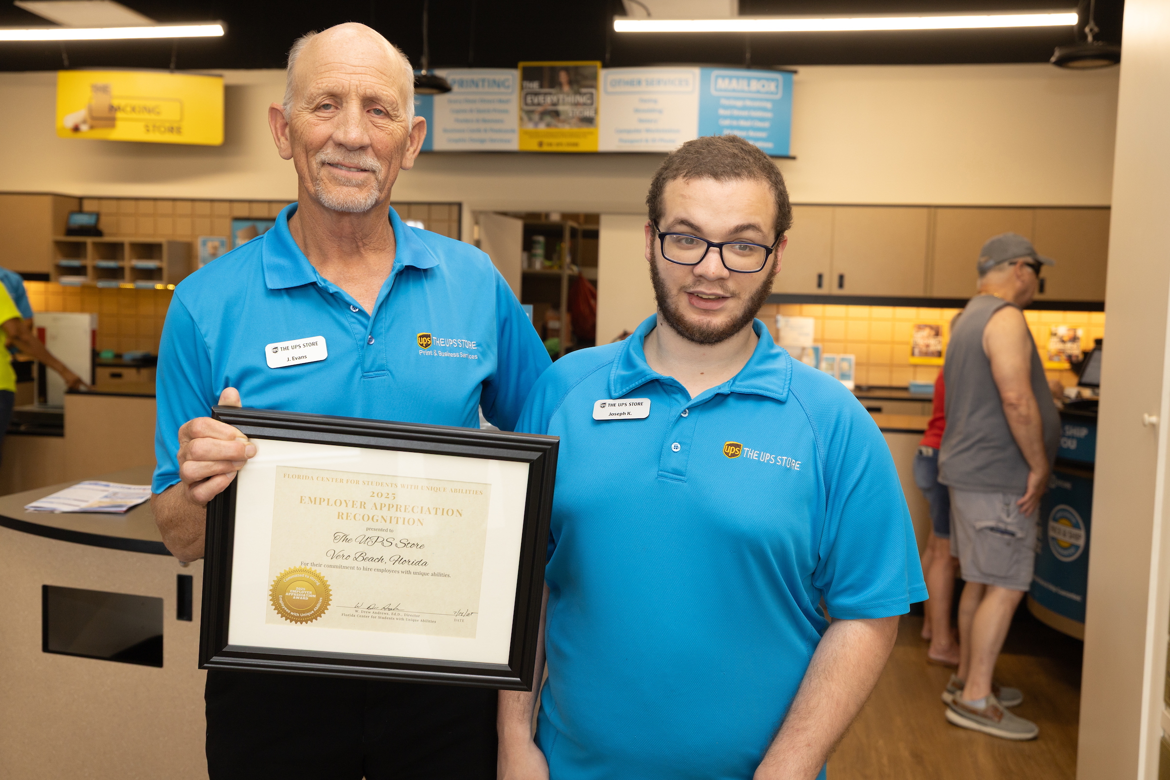 Two employees at The UPS Store in Vero Beach pose together, smiling, while holding an Employer Appreciation Recognition certificate from the Florida Center for Students with Unique Abilities. Both are wearing blue UPS Store shirts inside the store’s workspace.
