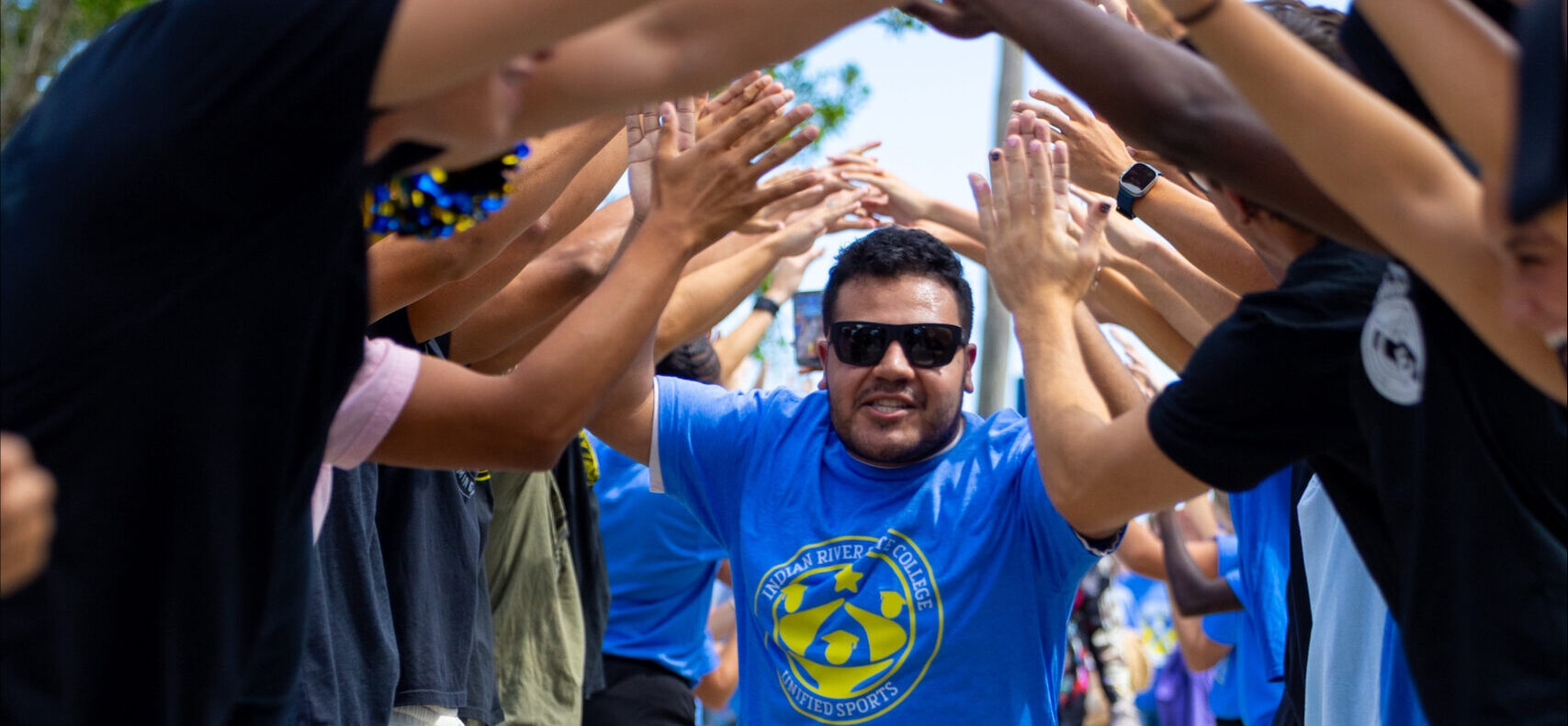 A man wearing sunglasses and an IRSC Unified Sports shirt runs through a tunnel formed by students raising their hands in celebration on a sunny campus walkway.